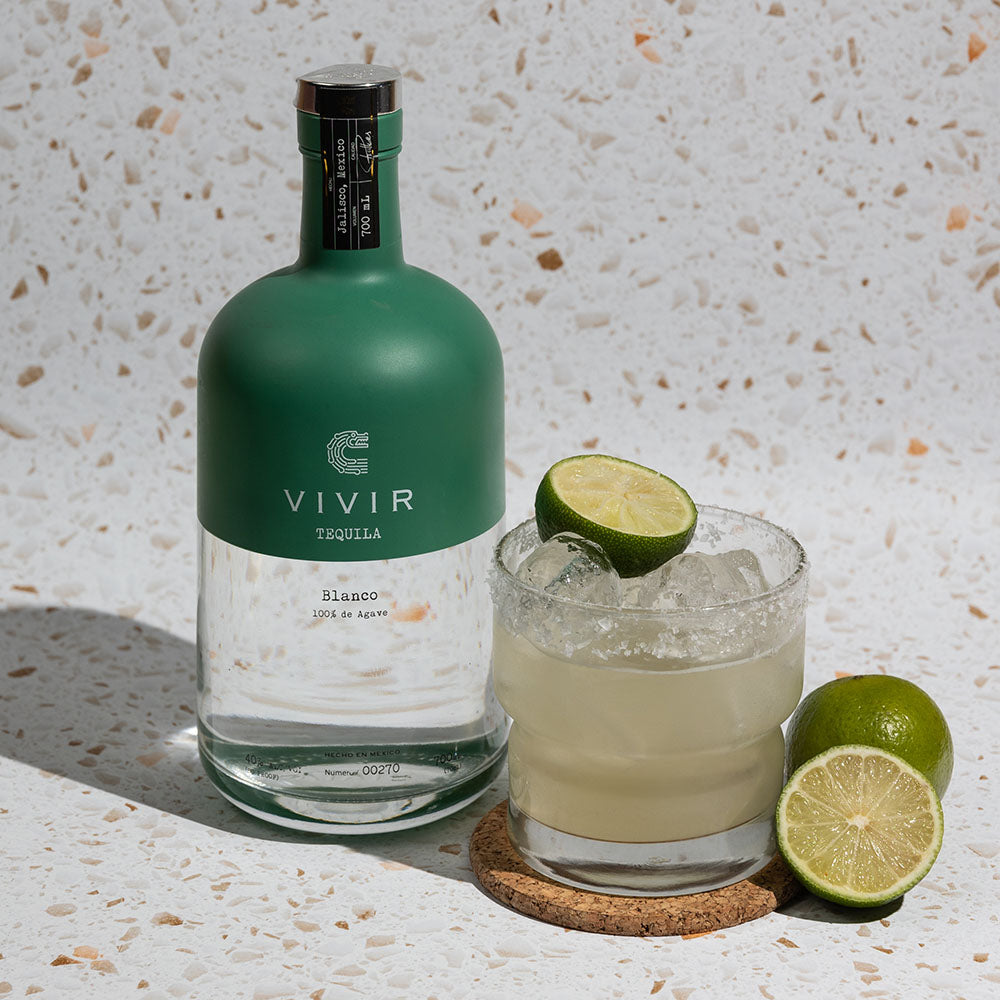 A bottle of VIVIR Tequila Blanco positioned next to a Margarita cocktail and some limes.