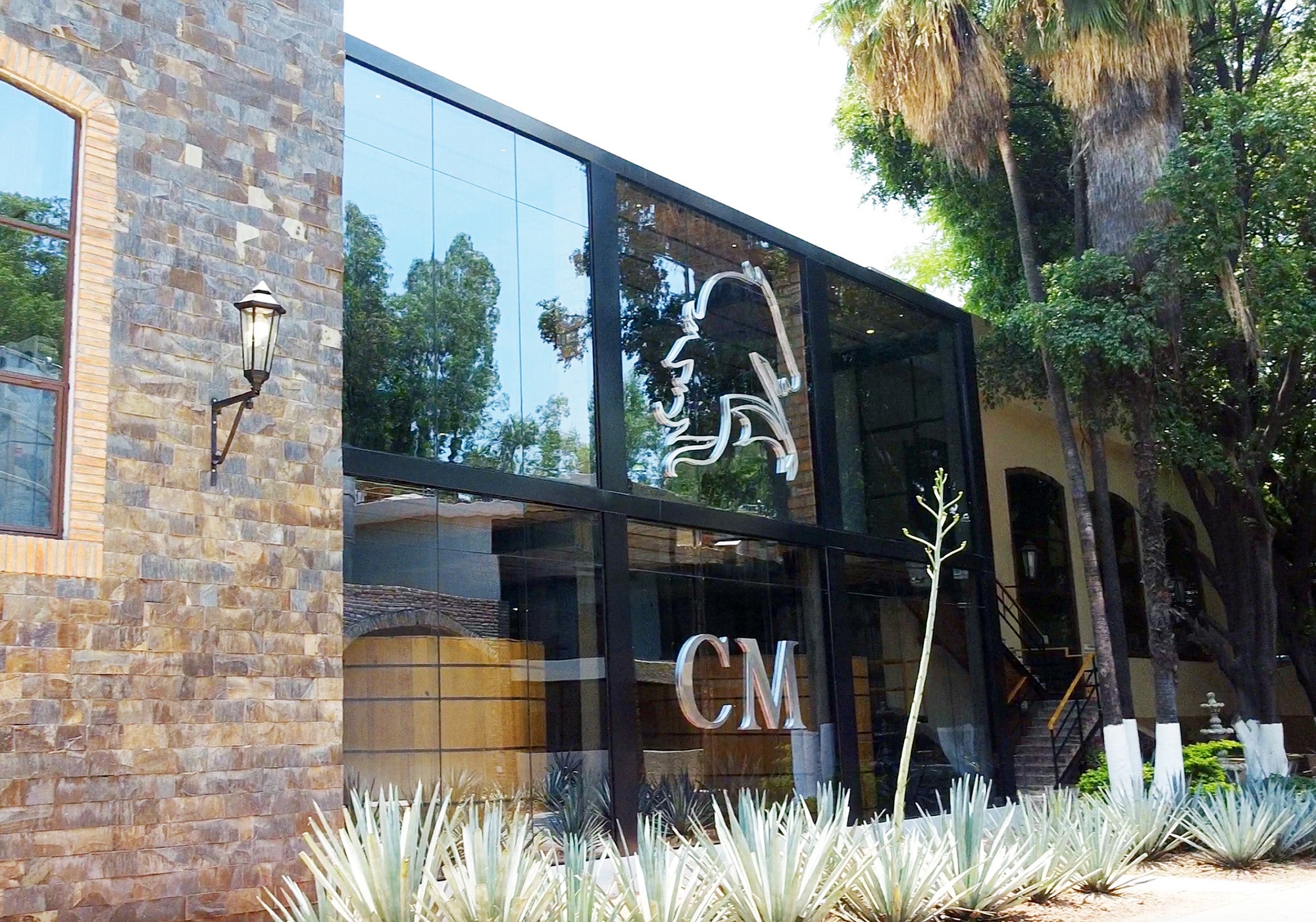 An exterior view of the glass walled tasting room at Casa Maestri distillery in Jalisco, Mexico where VIVIR Tequila is produced. Blue Weber Agave is seen in front of the Casa Maestri logo.