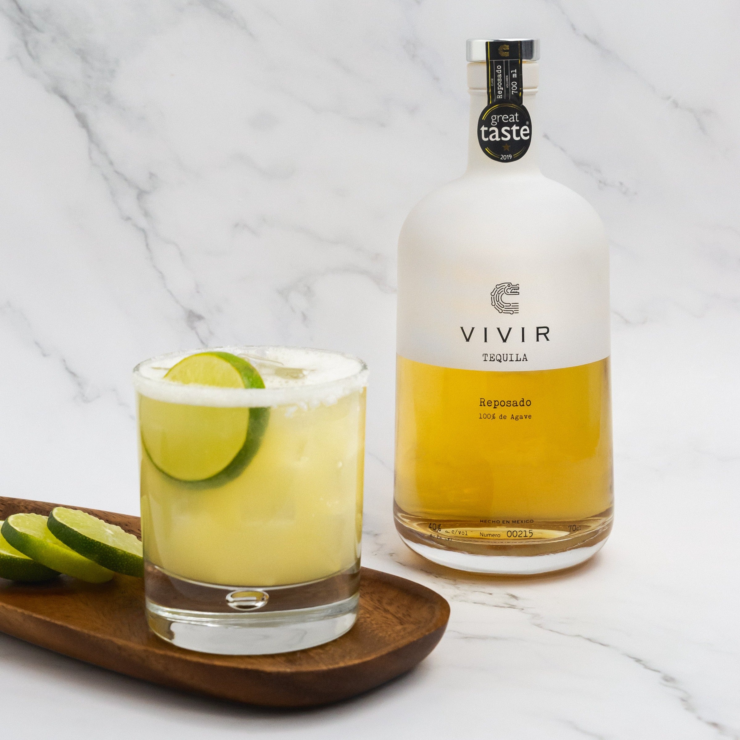 A bottle of VIVIR Tequila Reposado is positioned next to a Margarita cocktail in a tumbler glass with a lime wheel garnish.