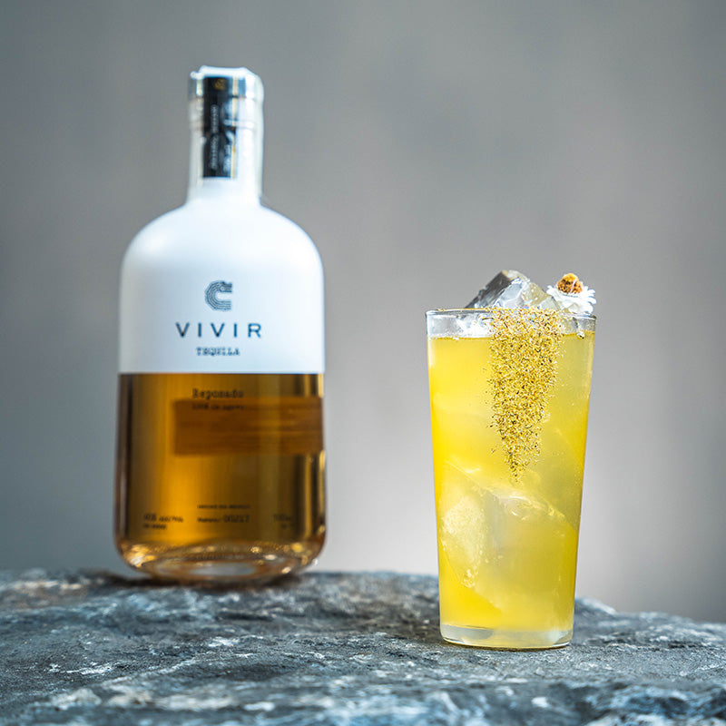 A bottle of VIVIR Tequila Reposado is positioned next to a yellow cocktail with a flower garnish in a highball glass.