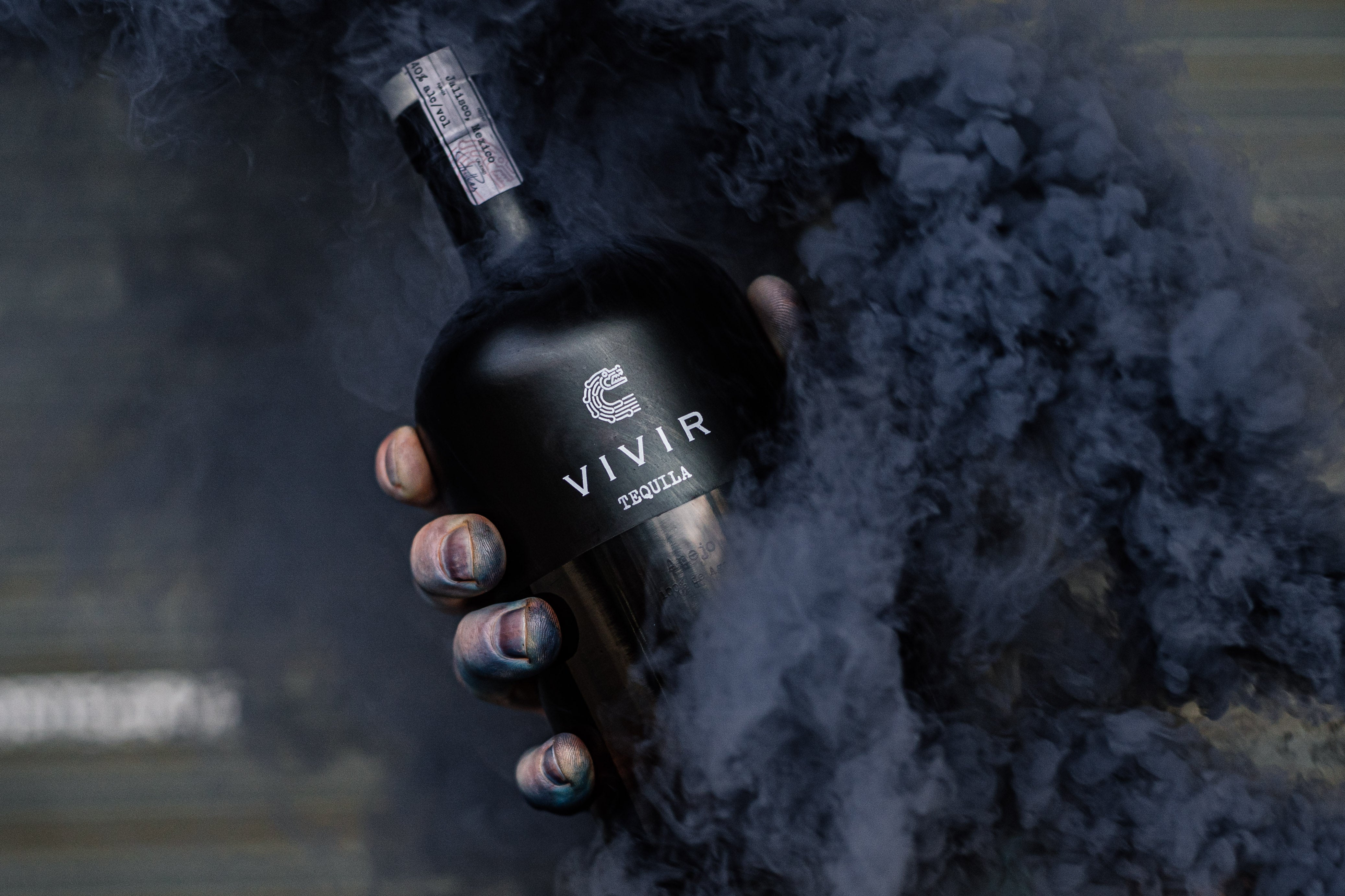A bottle of VIVIR Tequila Añejo in hand and appearing through navy black smoke.