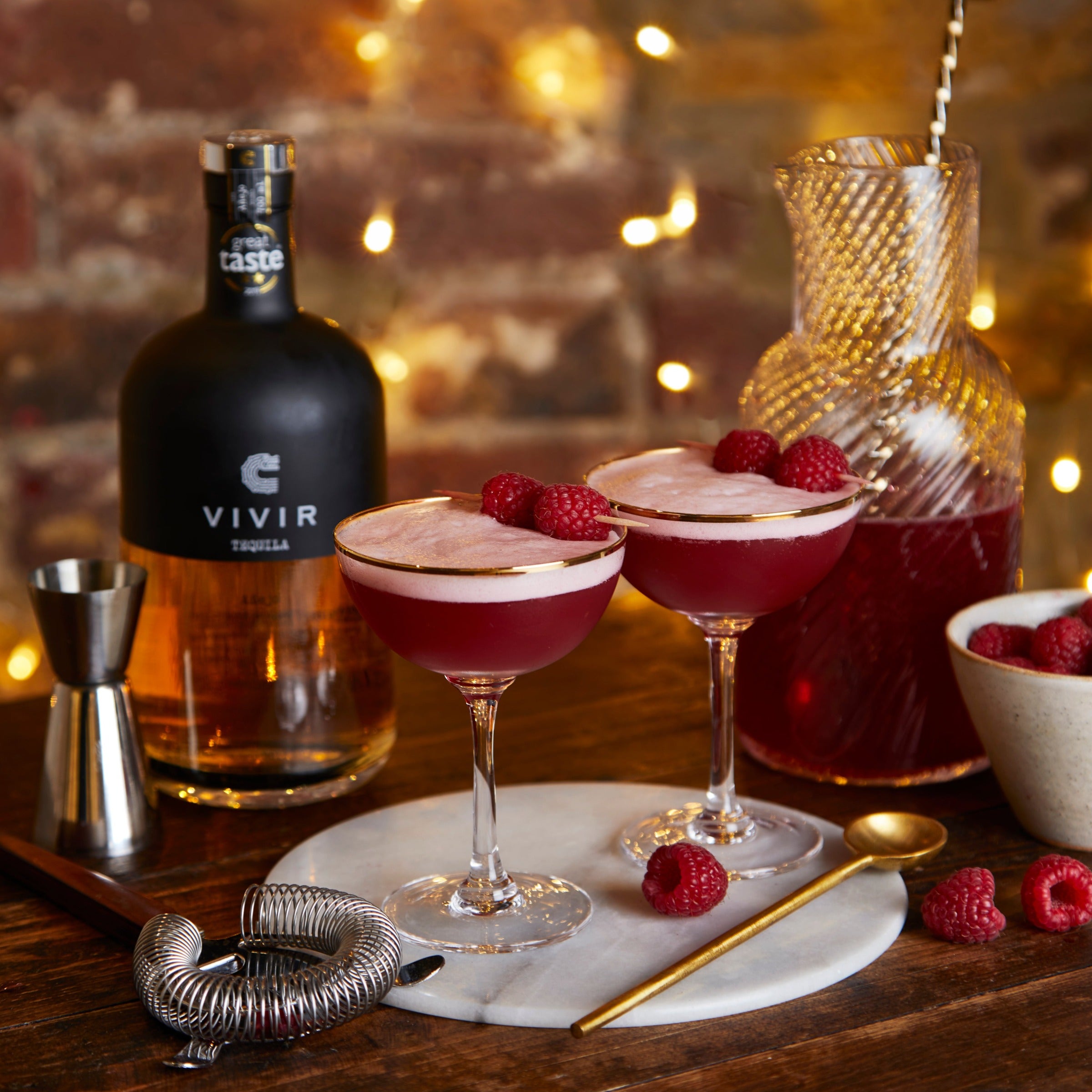 A bottle of VIVIR Tequila Añejo stands next to two rapsberry cocktails in martini glasses and a carafe containing the crimson cocktail, and surrounded by fresh raspberries and cocktail making equipment.